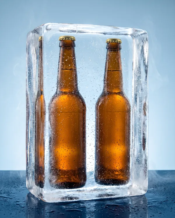 Tres botellas de cerveza ámbar con su corcholata atrapadas dentro de un bloque de hielo transparente, sobre fondo azul claro